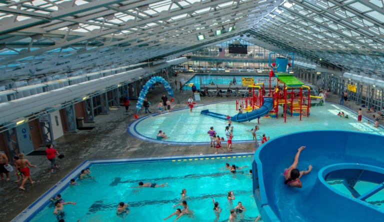 Swimmers play on a yellow, intertwined water slide inside a huge indoor pool with lots of natural light