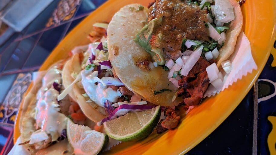 Tacos in fresh handmade tortillas piled high, on an orange plate with slices of lime