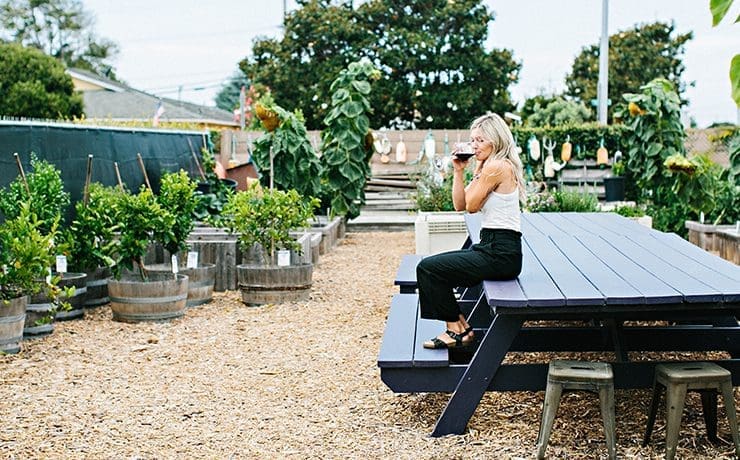 A woman sits on a purple picnic table, savoring Montemar Wines from her cup, surrounded by an outdoor garden adorned with potted plants and mulch ground.