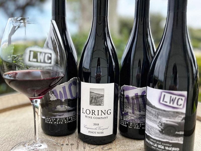 Four bottles of fine wine from Loring Wine Company, accompanied by a poured glass on a rustic wooden surface outdoors.