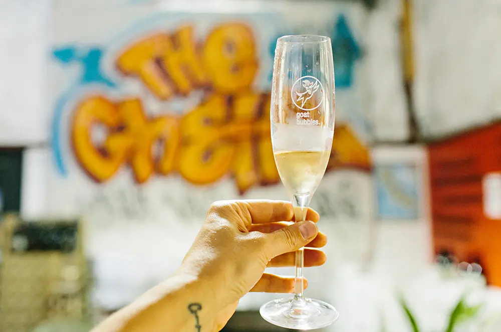 A champagne flute etched with a 'goat bubbles.' logo is held aloft, vignetted by a graffiti 'The Ghetto' mural