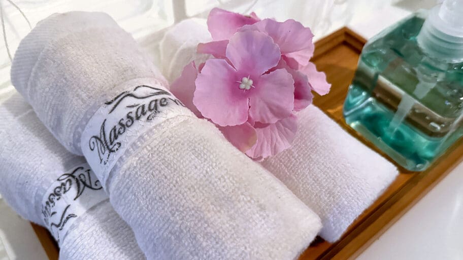 Rolled white towels and a pink flower rest elegantly on a wooden tray beside a pump bottle of liquid soap, creating the perfect spa ambiance.