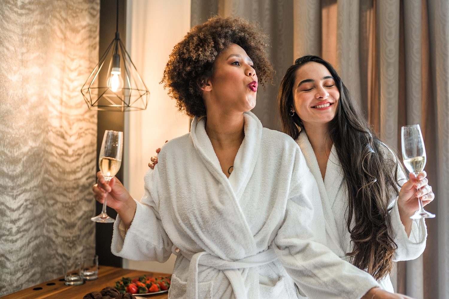Front view shot of two mixed race young women having fun in a hotel room, drinking their wine, eating chocolate muffins and enjoying their day.