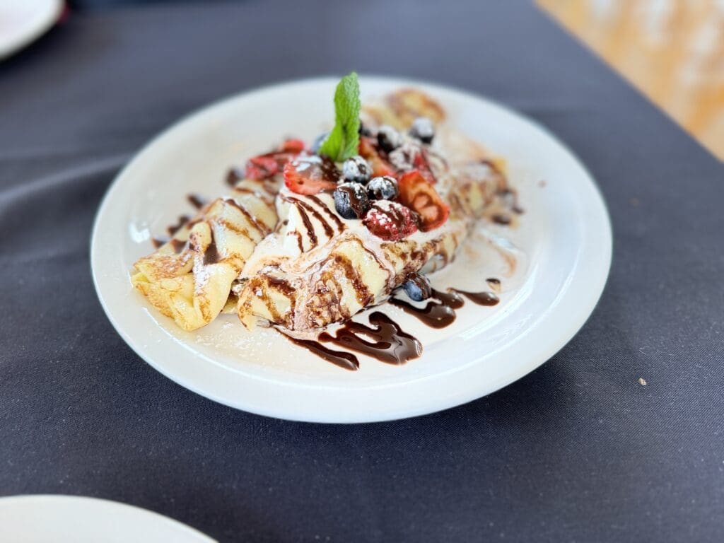 Crepes from Mamas Caffe topped with strawberries, blueberries, mint leaf, and a chocolate drizzle on a white plate set against a dark tablecloth background.