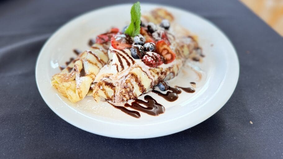 Crepes from Mamas Caffe topped with strawberries, blueberries, mint leaf, and a chocolate drizzle on a white plate set against a dark tablecloth background.