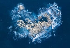 Aerial view of ocean waves crashing against a rocky formation, surrounded by deep blue water. This mesmerizing scene mirrors the marvels you can explore during the Cabrillo High School Aquarium Open House, where the mysteries of the ocean come to life.