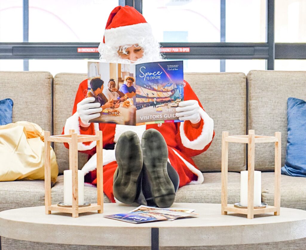 Santa Claus reads the Ultimate Holiday Guide while sitting on a sofa in Lompoc, with a gift bag beside him and wooden decor on the table.