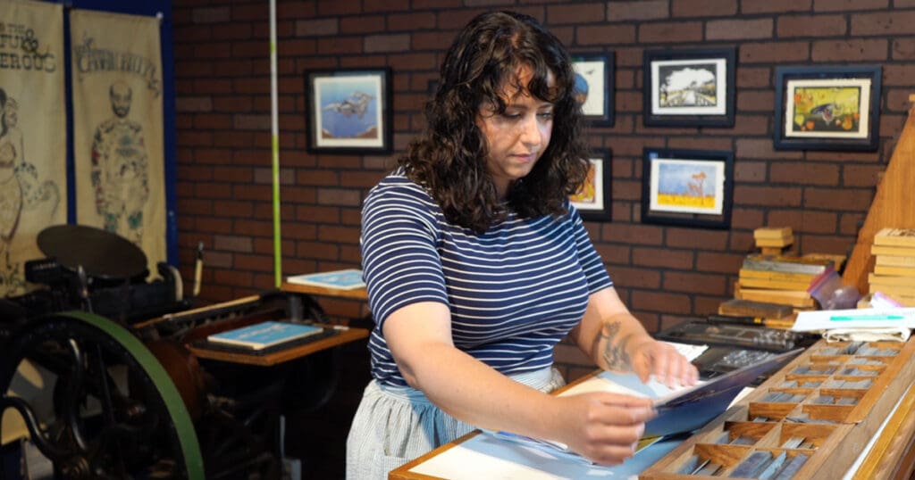 At the printing studio, Angelina LaPointe meticulously arranges papers, her focus undisturbed by the vibrant artwork adorning the brick wall behind her. The pieces, reminiscent of the California Wilds, await their moment to shine at her upcoming artist reception.