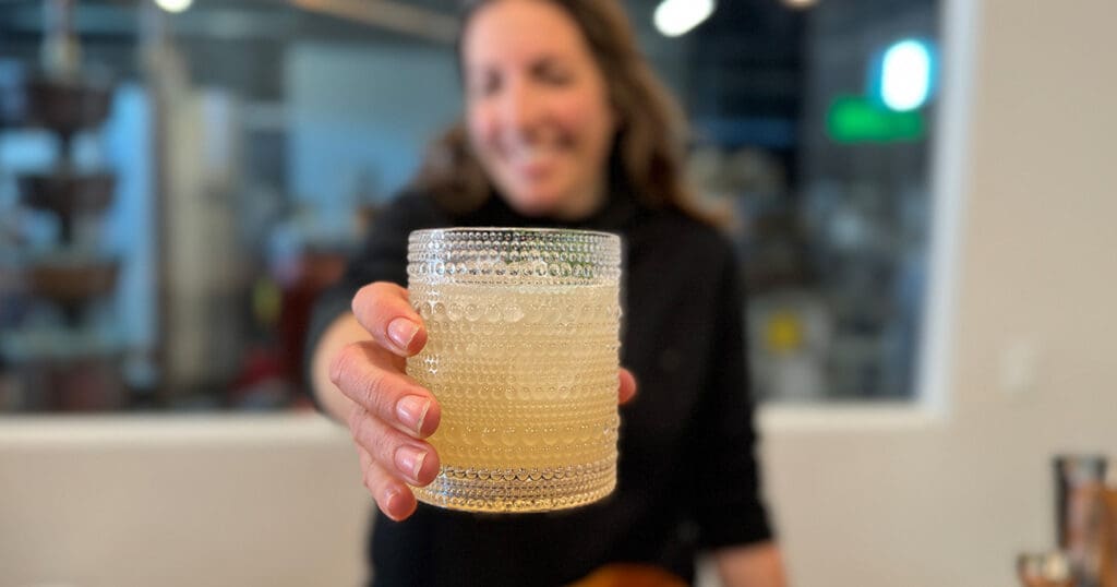 A person is holding a decorative glass filled with a light-colored cocktail, extending it towards the camera. The background is elegantly blurred, adding an air of class to the scene.