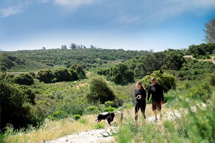 Two people walking a dog on a trail through a lush green landscape in Lompoc, CA, under a partly cloudy sky.