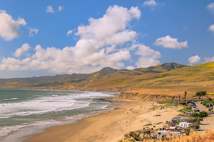 A scenic coastline near Lompoc, CA, with waves lapping onto the sandy beach, flanked by green hills under a partly cloudy blue sky. In the distance, a few cars and people can be seen enjoying the serene landscape.