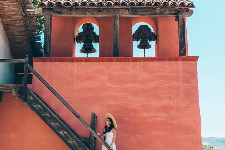 A woman in a straw hat ascends the stairs next to a red-orange building with two bells under arches on the roof, capturing the quaint charm of Lompoc, CA.