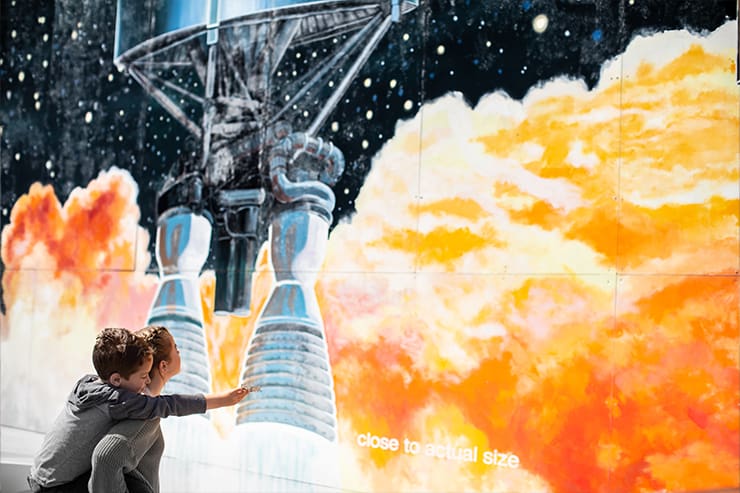 A child and an adult gaze at a mural in Lompoc, CA, depicting a rocket launch with vivid orange flames and billowing smoke. Text reads close to actual size.