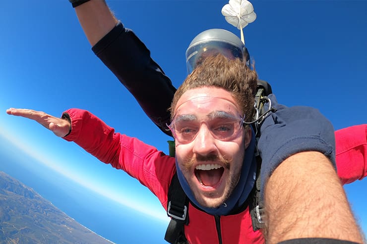 Soaring through the clear blue sky above Lompoc, CA, a person in a red suit and goggles experiences the thrill of tandem skydiving, arms outstretched as they take in the breathtaking landscape below.