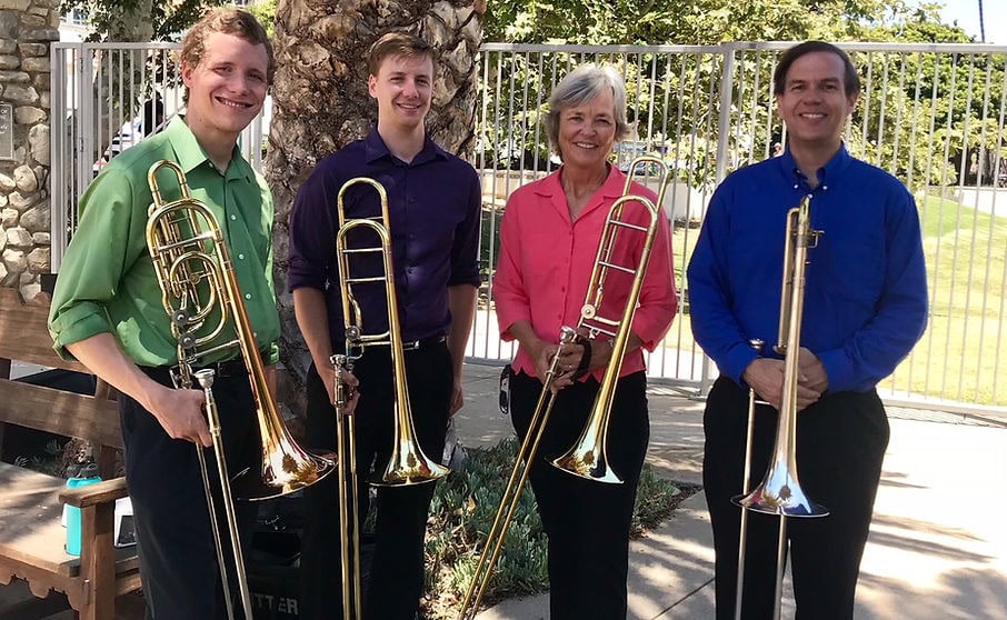 Four people from the Slideways Trombone Quartet stand outdoors holding trombones, smiling at the camera. Dressed in brightly colored shirts, they pose in front of a metal fence and trees before their Free Outdoor Concert.