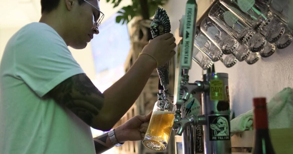 A person in a white shirt pours Lompoc brews into a glass mug from a tap at a bar, with several other mugs hanging on hooks above—perfect for those seeking late-night bites and drinks.