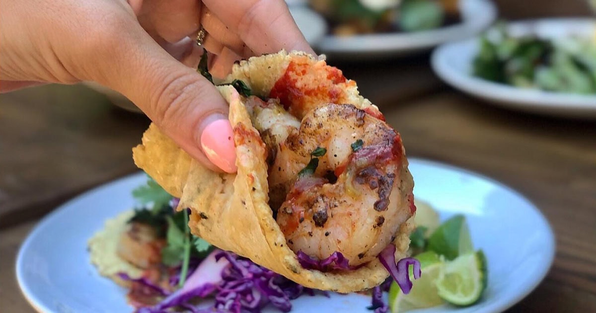 A hand holds a shrimp taco filled with grilled shrimp, shredded purple cabbage, and cilantro, with plates of food and lime wedges in the background—just the kind of bite Cisko Kid Eats raves about in his food reviews.