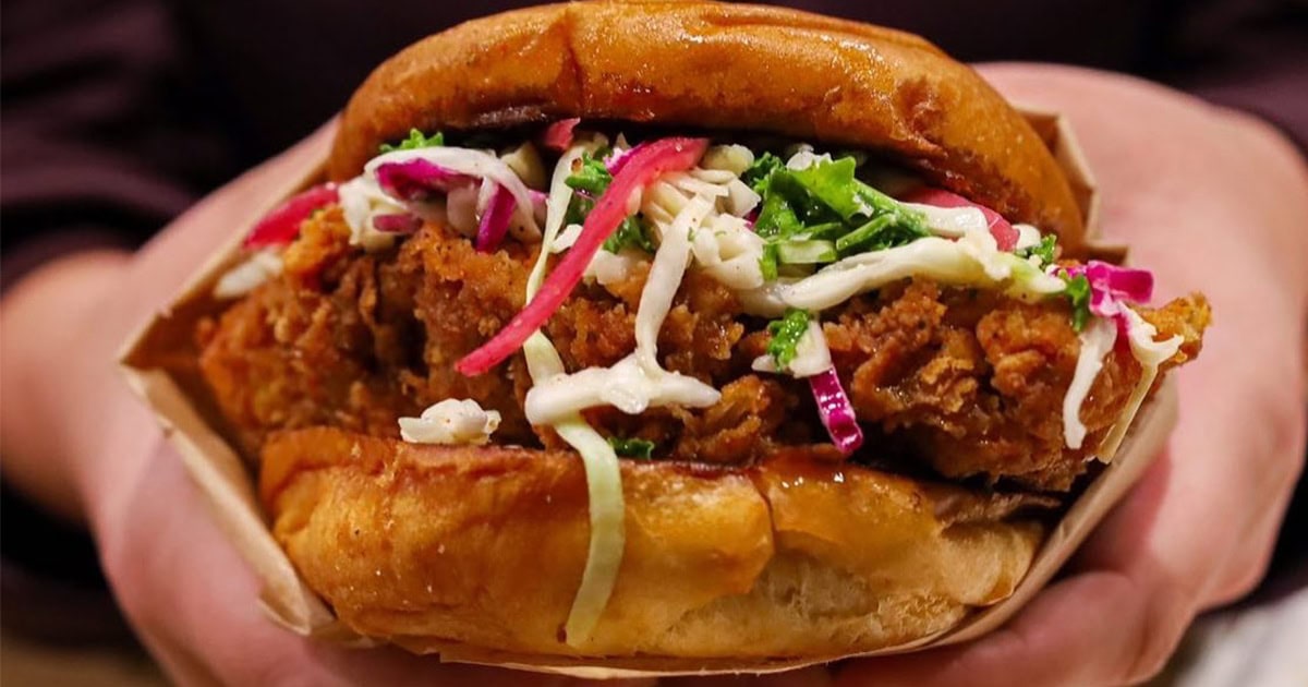 A close-up of hands holding a fried chicken sandwich topped with coleslaw and served on a toasted bun.